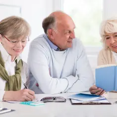 Trzech seniorów przy stole, jeden pisze, drugi czyta książkę. Wesołe zajęcia dla seniorów.