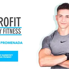 Uśmiechnięty, muskularny zdrofit trener personalny w szarej koszulce, z logo ZDROFIT KLUBY FITNESS i informacją o darmowym treningu próbnym.
