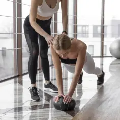 Trening siłowy i cardio na redukcji. Kobieta wykonuje pompki z piłką, druga osoba ją asekuruje.