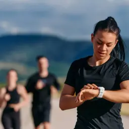 Kobieta sprawdza zegarek podczas biegania. Grupa biegaczy w tle, być może trening w stylu bieganie metodą Danielsa.