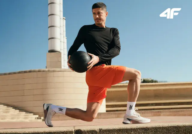 Robert Lewandowski w pomarańczowych spodenkach i czarnej bluzie wykonuje ćwiczenie z piłką. Idealne jakie buty do crossfitu zapewniają stabilność.